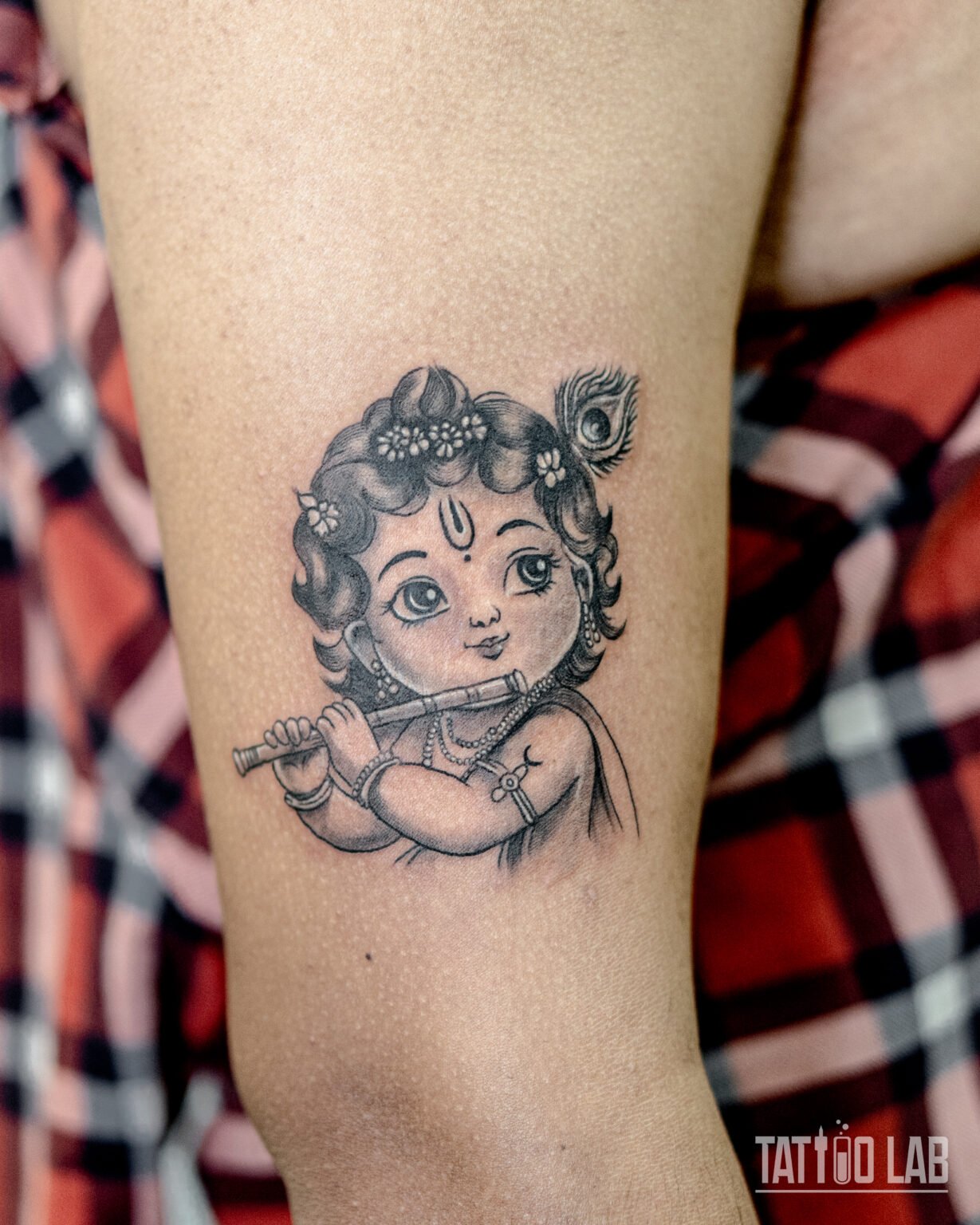 Black and grey baby Krishna tattoo playing the flute with a peacock feather, on the back of the arm, Tattoo Lab, Halifax