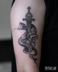 Black and grey tattoo of a lion-hilt dagger through an anatomical heart with a coiled snake on the upper arm