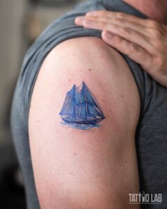 Small blue Bluenose-style schooner tattoo on the upper arm, a Nova Scotia maritime design in fine line colour