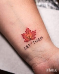 Red and orange maple leaf tattoo above the words "LET THEM" on the wrist, small colour realism with fine lettering