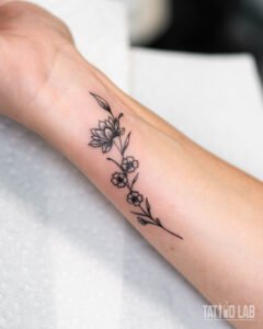 Fine line wrist tattoo of a lotus flower and small blossoms on a slender branch, delicate black linework