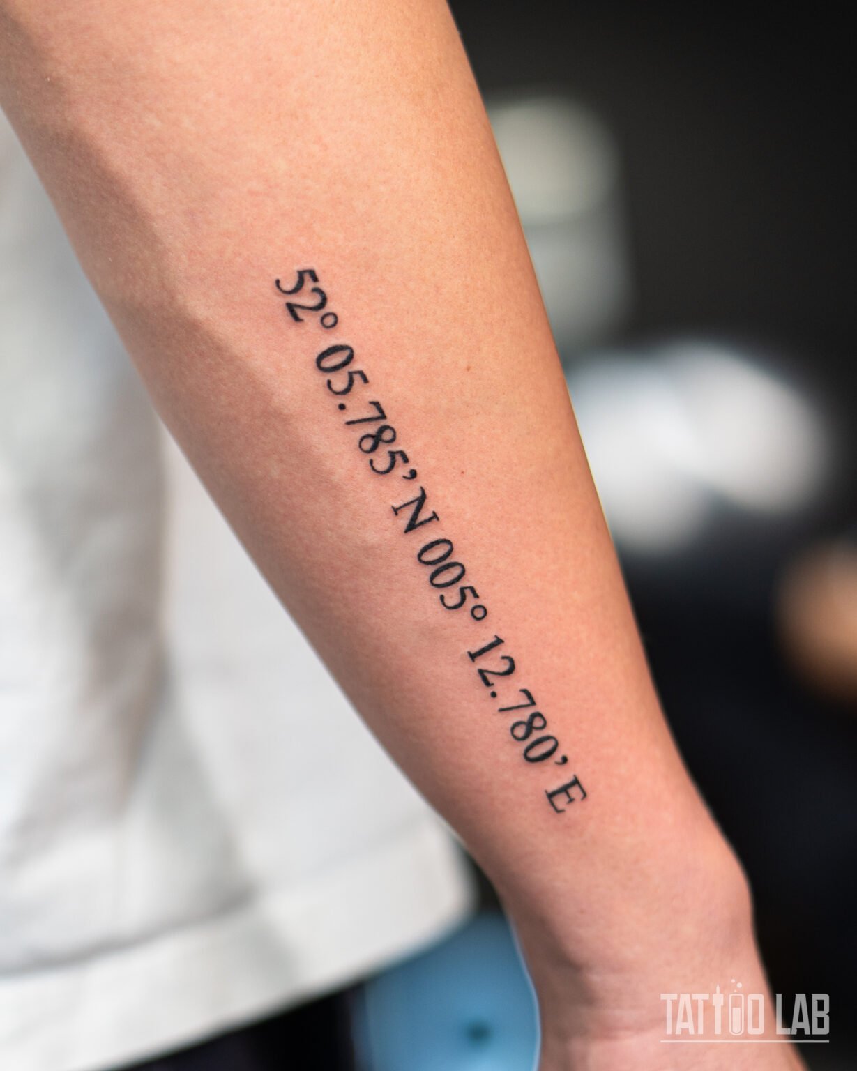 Minimalist GPS coordinates tattoo in black serif numerals running along the forearm, a clean lettering design, Tattoo Lab, Halifax