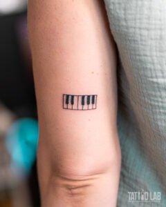 Small black ink piano keys tattoo on the upper arm, a minimalist design for music lovers