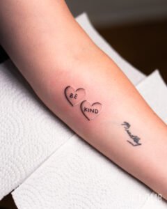 Two small shaded heart tattoos reading "BE" and "KIND" on the forearm, a minimalist black ink design