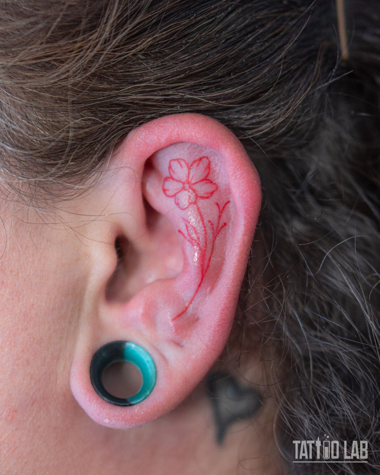 Small red ink fine line flower tattoo inside the upper ear, a delicate minimalist design with a slender stem and leaves, Tattoo Lab, Halifax