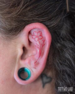 Small red ink fine line flower tattoo inside the upper ear, a delicate minimalist design with a slender stem and leaves