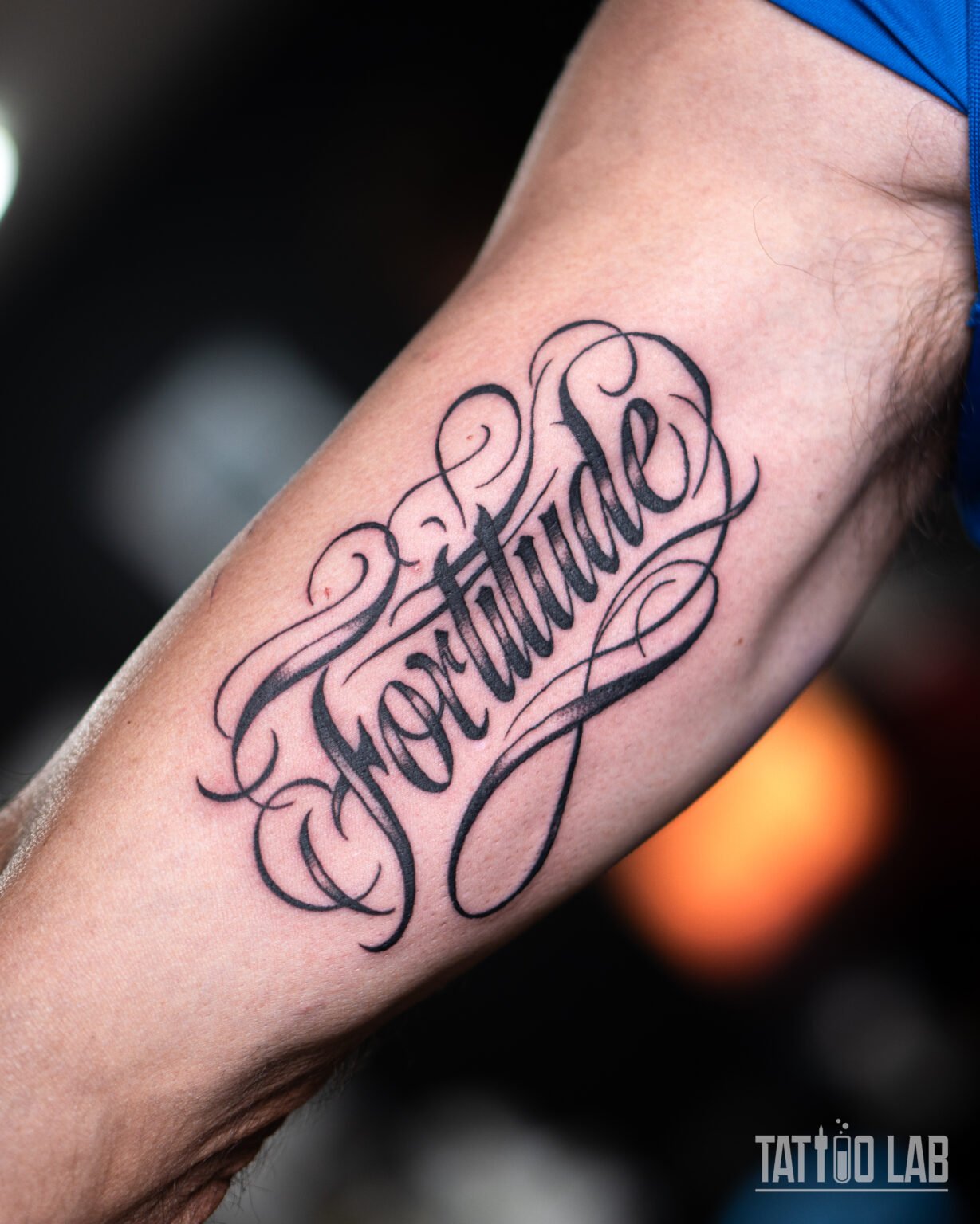 Black script lettering tattoo reading "Fortitude" with flowing flourishes on the inner upper arm, Tattoo Lab, Halifax