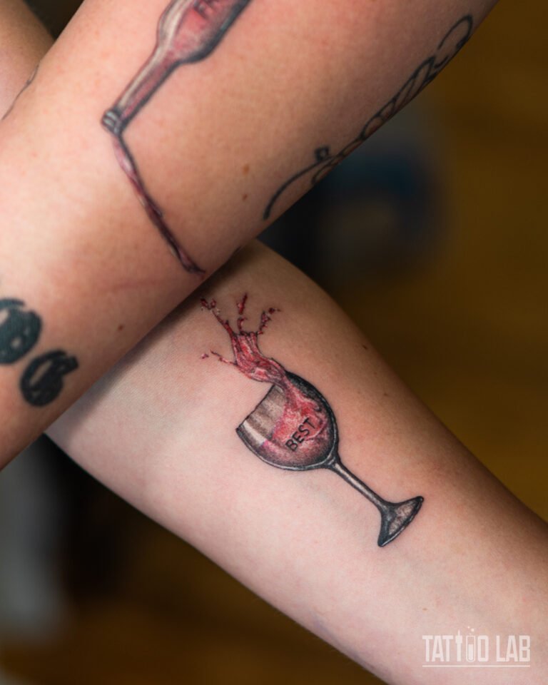 Wine glass tattoo labelled "BEST" with a red wine splash on the forearm, half of a matching best friends set