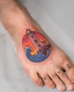 Colour lighthouse tattoo on the top of the foot with a crashing wave and orange sunset sky