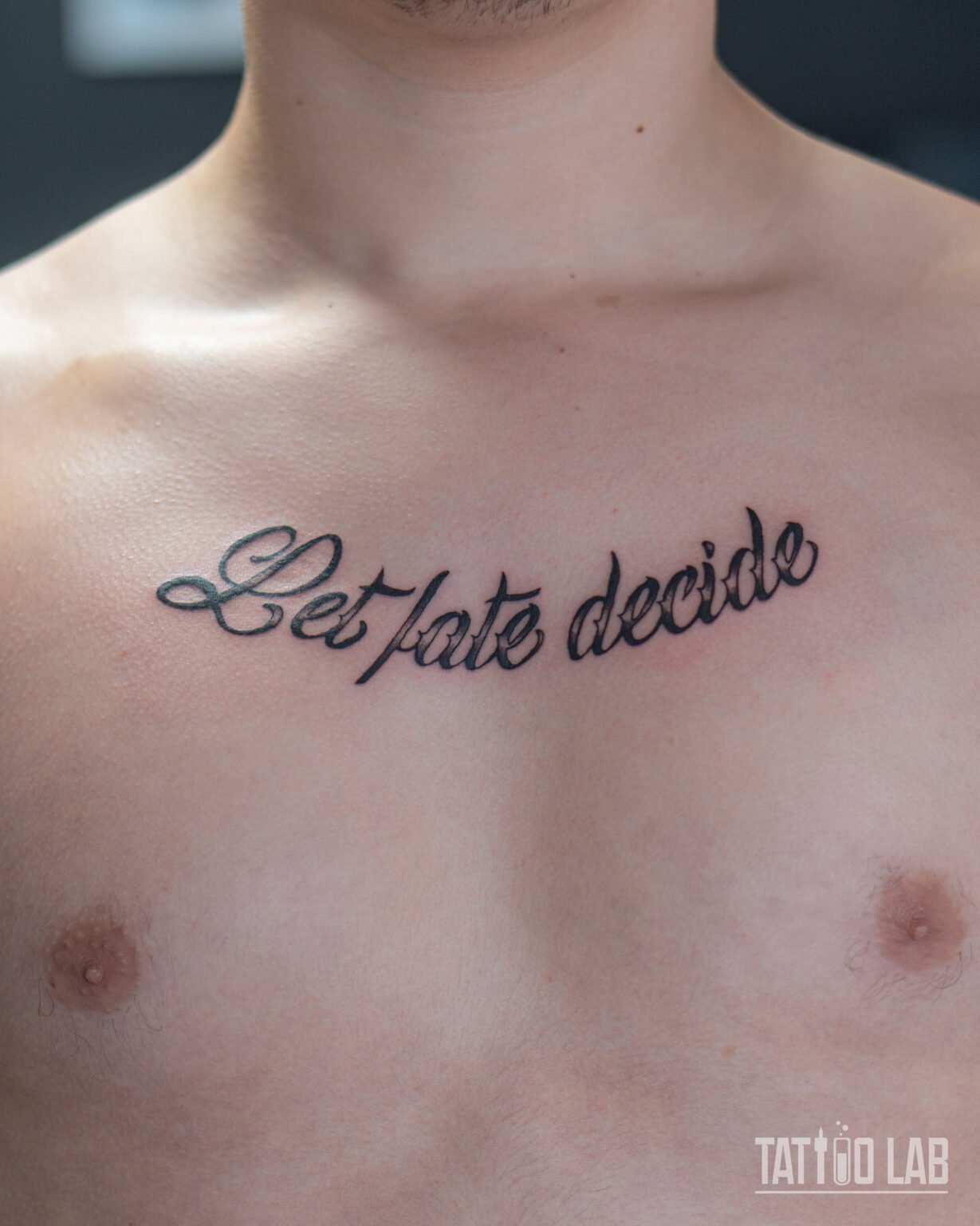 Script lettering tattoo reading "Let fate decide" across the upper chest in bold black cursive, Tattoo Lab, Halifax