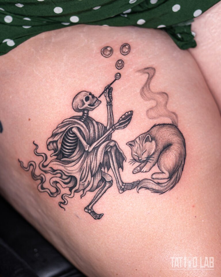 Black and grey thigh tattoo of a robed skeleton blowing bubbles beside a sleeping cat