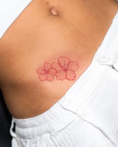 Two red ink hibiscus flowers tattoo on the hip, a delicate fine line floral design with soft colour shading