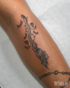 Witch broom tattoo with climbing vines, bats and crescent moons on the forearm in fine line black ink