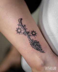 Fine line witch broom tattoo with two suns, tiny flowers and dotwork on the lower forearm