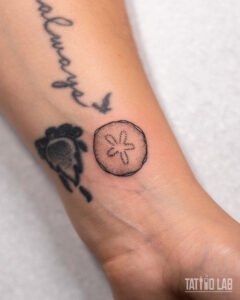Small sand dollar tattoo on the wrist with a fine outline and stipple shading, a beach-inspired design