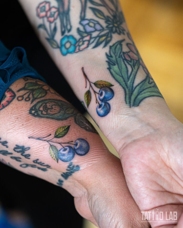 Matching colour realism blueberry tattoos on two wrists, small blue berries on a stem with green leaves