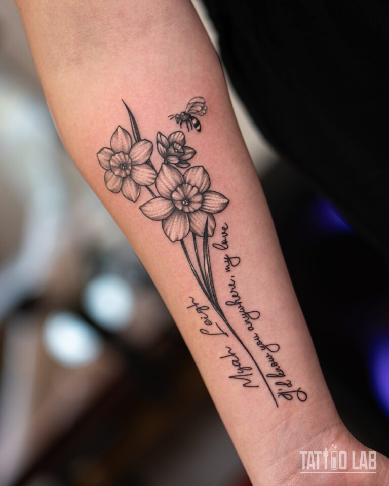 Fine line forearm tattoo of daffodils and a bee with a handwritten script line along the stem, black and grey