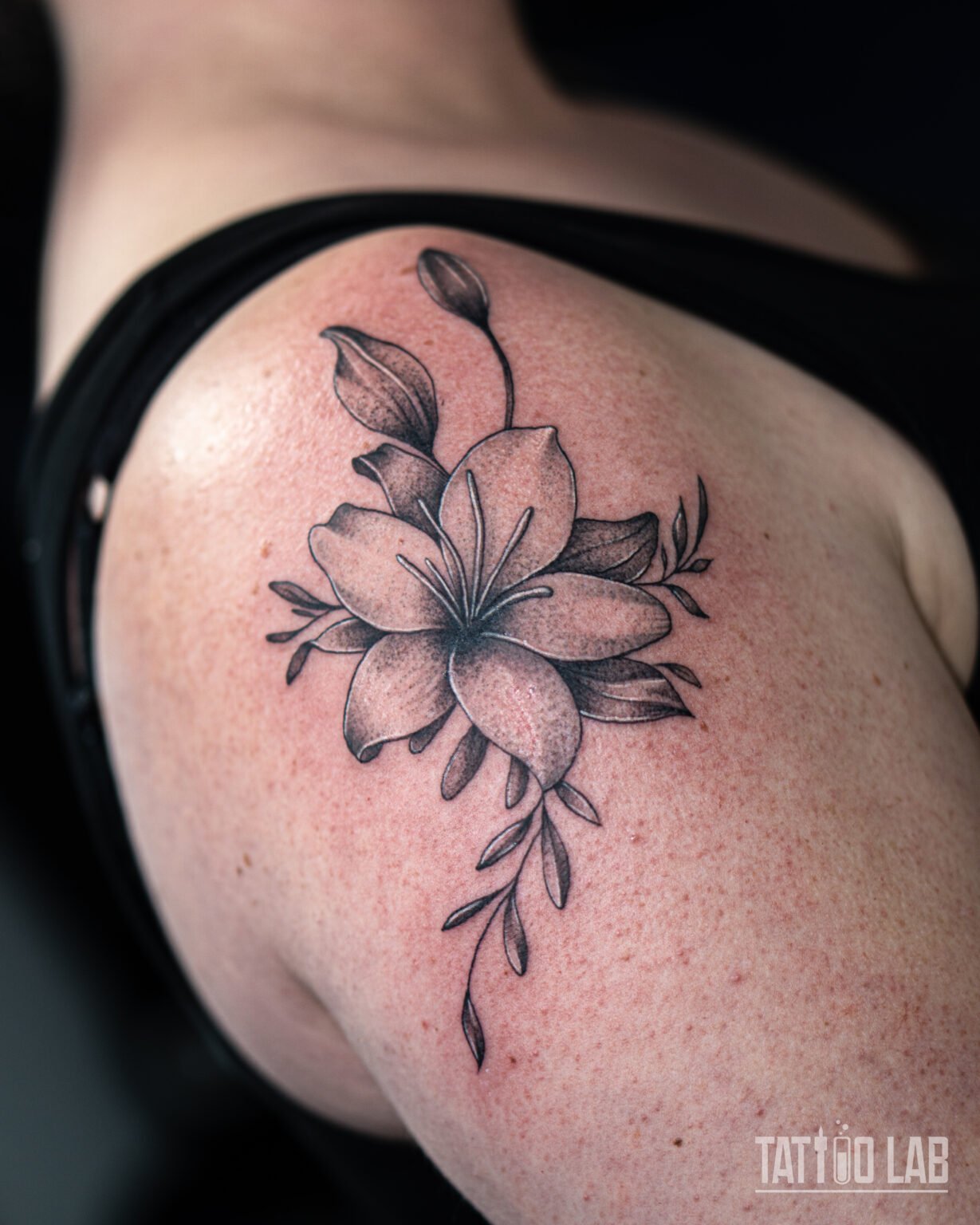 Black and grey lily tattoo on the shoulder with a flower bud, leaves and soft stipple shading, Tattoo Lab, Halifax