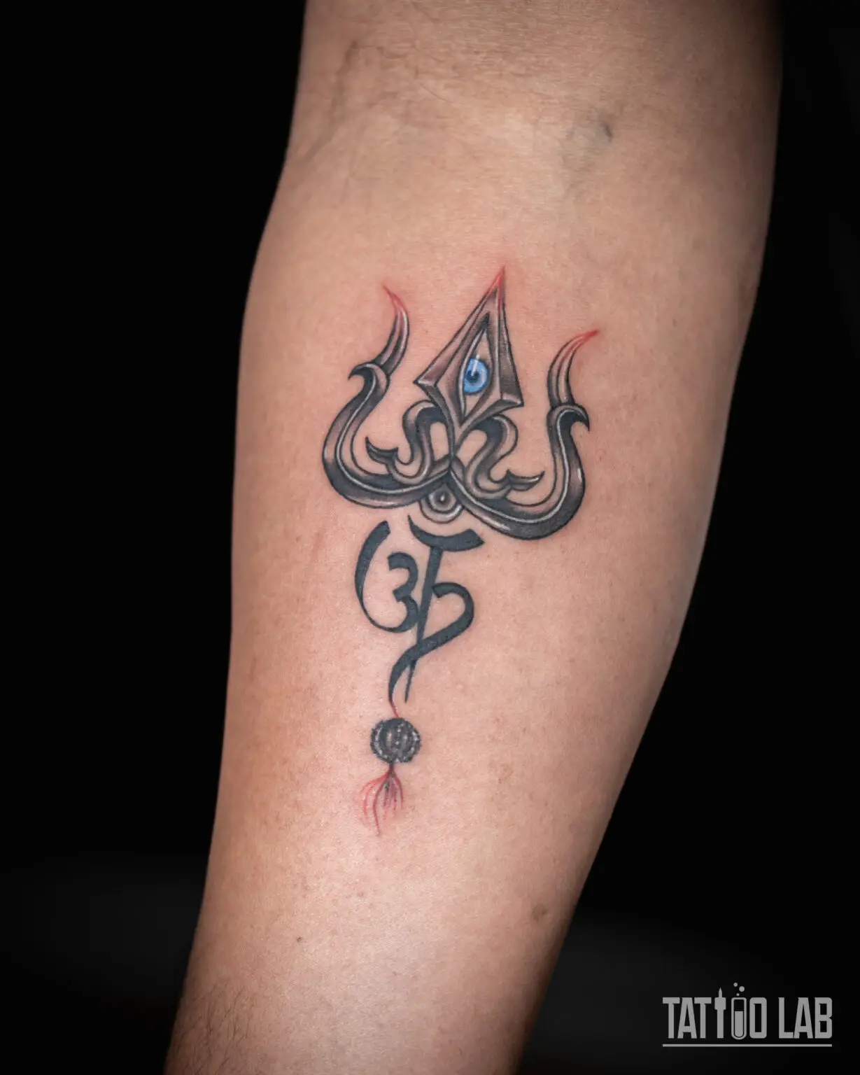 Trishul and Om symbol tattoo on the forearm with a blue eye, hanging bead and fine red accents, Tattoo Lab, Halifax