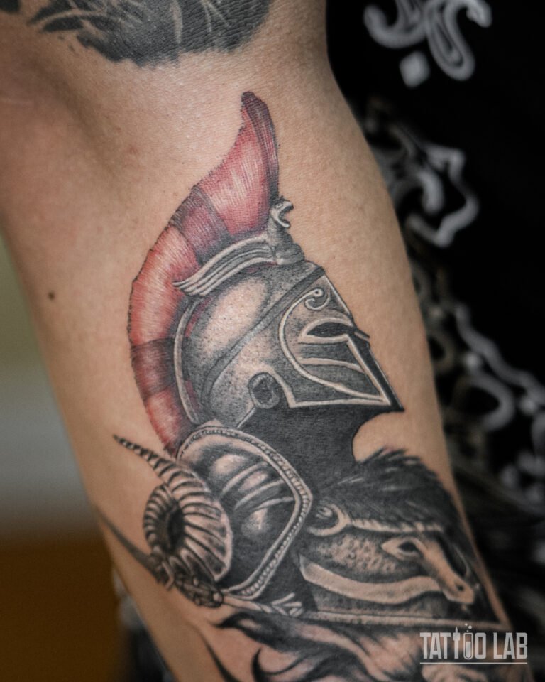 Spartan helmet tattoo with a red plume and ram horn detail, black and grey with colour accents, on the arm