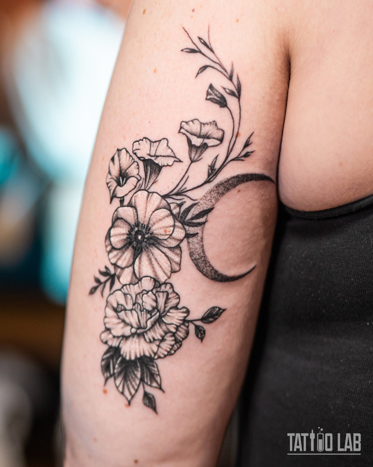 Black and grey floral tattoo with blooming flowers and a dotwork crescent moon on the back of the upper arm, Tattoo Lab, Halifax