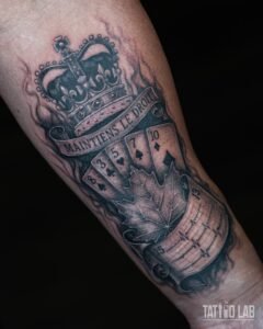 Crown, banner, playing cards, maple leaf and heartbeat scroll tattoo in black and grey realism on forearm