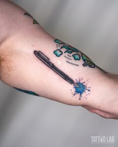 Black and grey realism fountain pen tattoo with a blue watercolour ink splash at the nib on the inner upper arm