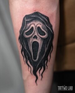 Black and grey horror movie ghost mask tattoo with a flowing solid black hood on the forearm