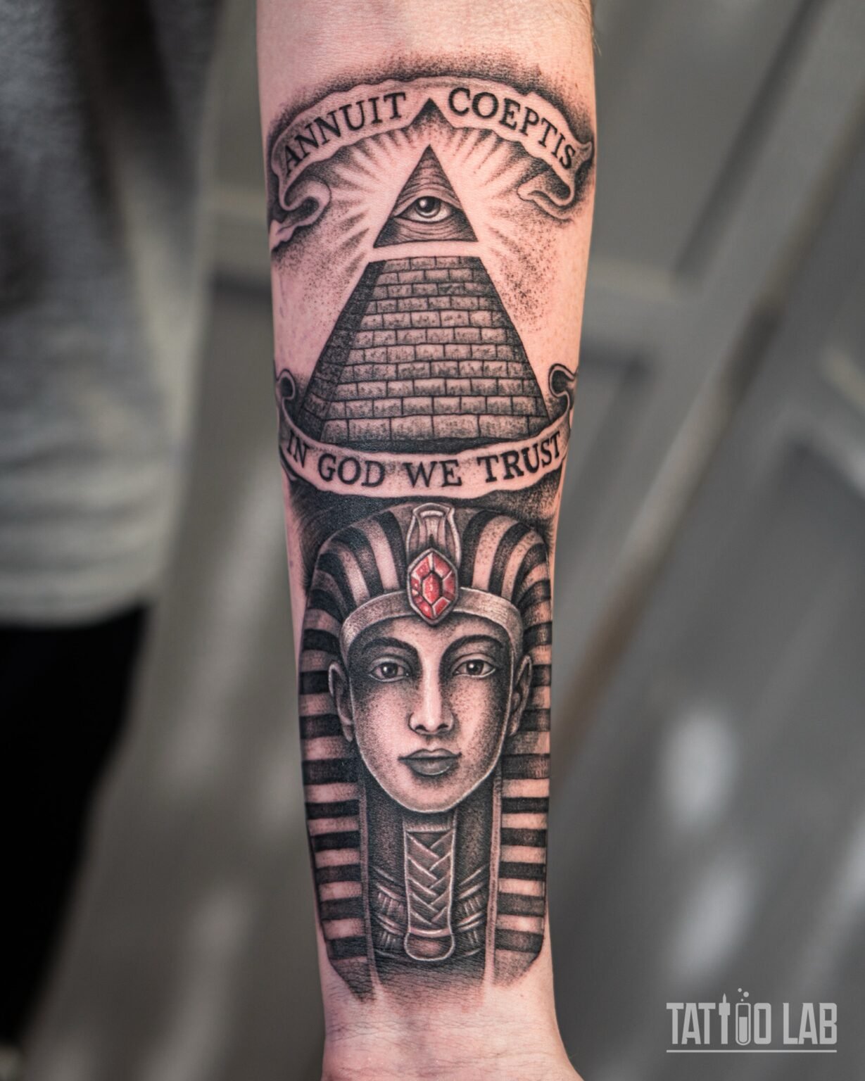Black and grey realism pharaoh tattoo with a red gem, below an all-seeing eye pyramid and banners, on the forearm, Tattoo Lab, Halifax
