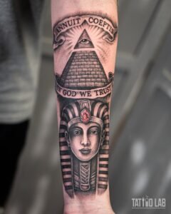 Black and grey realism pharaoh tattoo with a red gem, below an all-seeing eye pyramid and banners, on the forearm