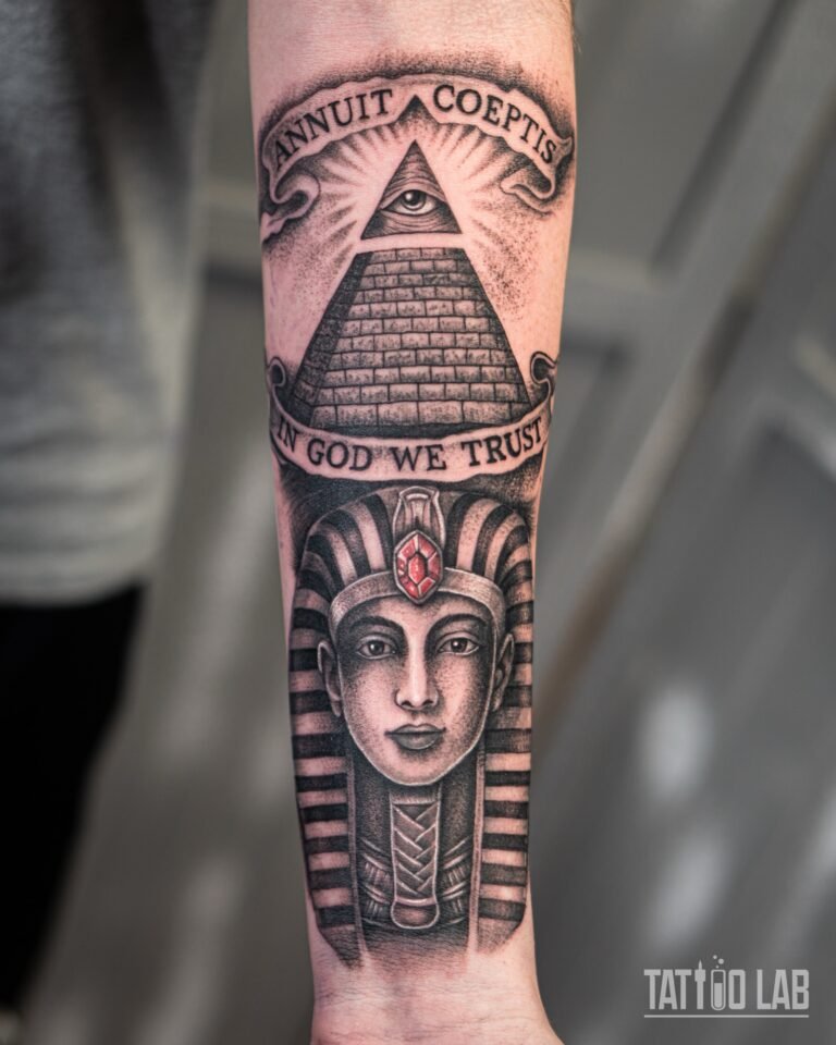 Black and grey realism pharaoh tattoo with a red gem, below an all-seeing eye pyramid and banners, on the forearm