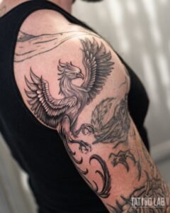 Black and grey realism phoenix tattoo with raised wings and curling tail feathers on the shoulder and upper arm