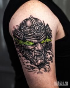 Black and grey realism tattoo of a samurai helmet and oni mask with glowing green eyes on the upper arm