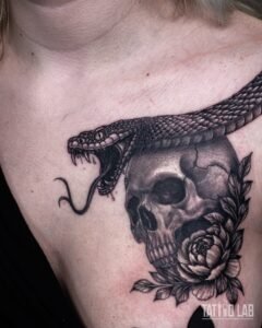 Black and grey realism tattoo of a striking snake over a cracked skull with a peony and leaves on the chest