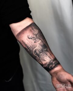 Black and grey realism tattoo of a stag standing in a misty pine forest on the inner forearm