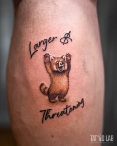 Colour cartoon red panda tattoo with raised paws and script lettering above and below, back of calf close-up