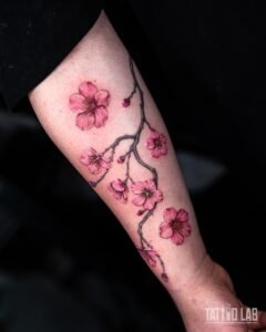 Pink cherry blossom branch tattoo in soft colour with grey shaded branches and buds along the inner forearm