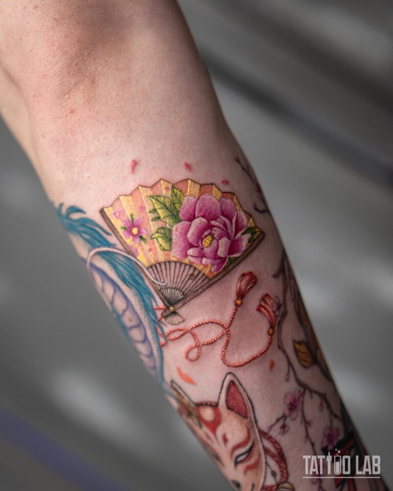 Colour tattoo of a Japanese folding fan with a pink peony and tassels on a forearm sleeve