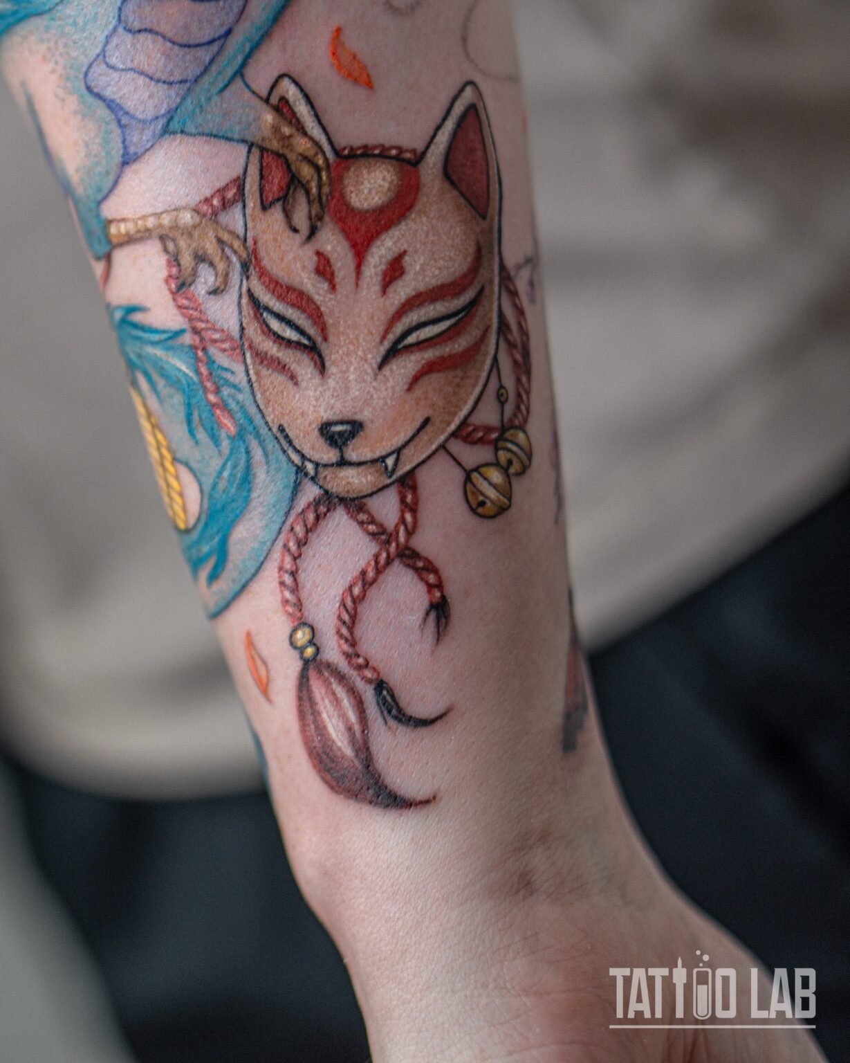 Colour kitsune fox mask tattoo with red rope, bells and tassel held by a dragon claw, part of a forearm sleeve, Tattoo Lab, Halifax