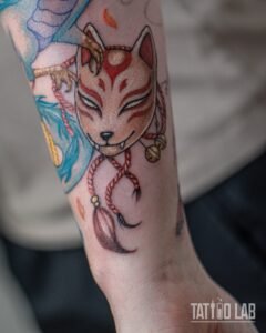 Colour kitsune fox mask tattoo with red rope, bells and tassel held by a dragon claw, part of a forearm sleeve