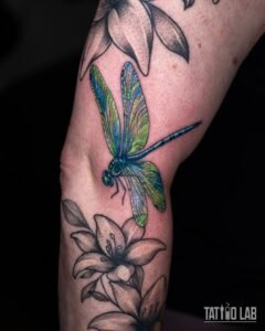 Colour realism dragonfly tattoo with iridescent green and blue wings between black and grey lily flowers, on the arm near the elbow