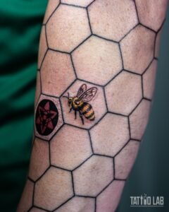 Realistic colour honeybee tattoo in a black honeycomb grid beside a red anime-style emblem on the forearm