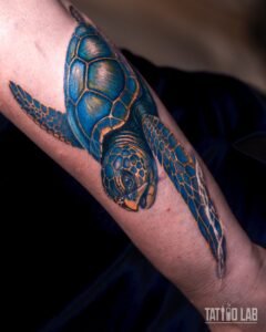 Sea turtle tattoo in blue and yellow-orange colour with a glossy shaded shell on forearm