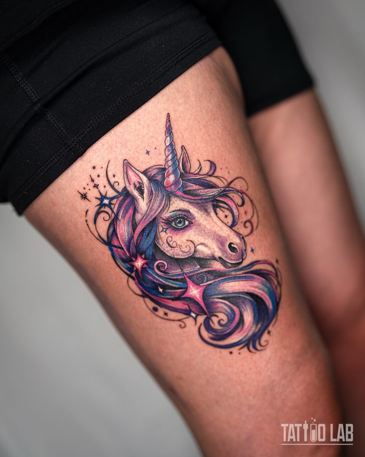 Colour unicorn head tattoo with a flowing pink, purple and blue mane and sparkles on the upper thigh, shown in a wider view, Tattoo Lab, Halifax