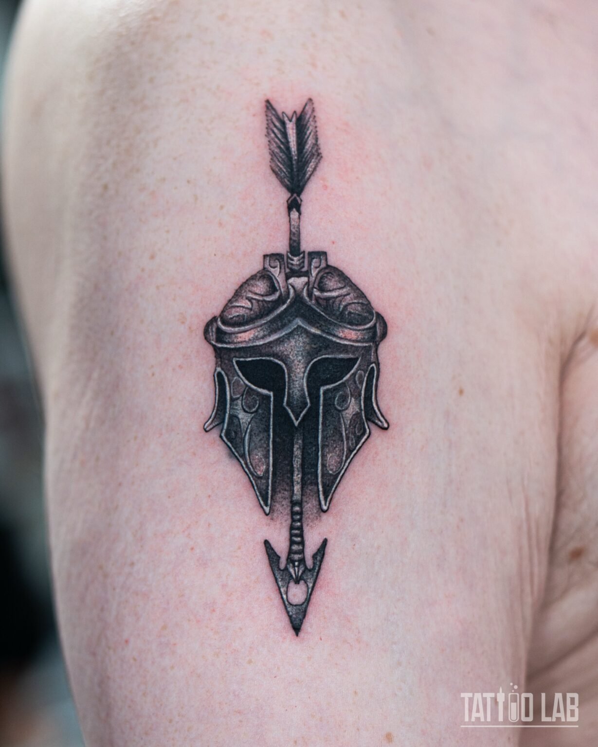 Black and grey cover-up tattoo of a Spartan helmet pierced by an arrow, on the upper arm, Tattoo Lab, Halifax