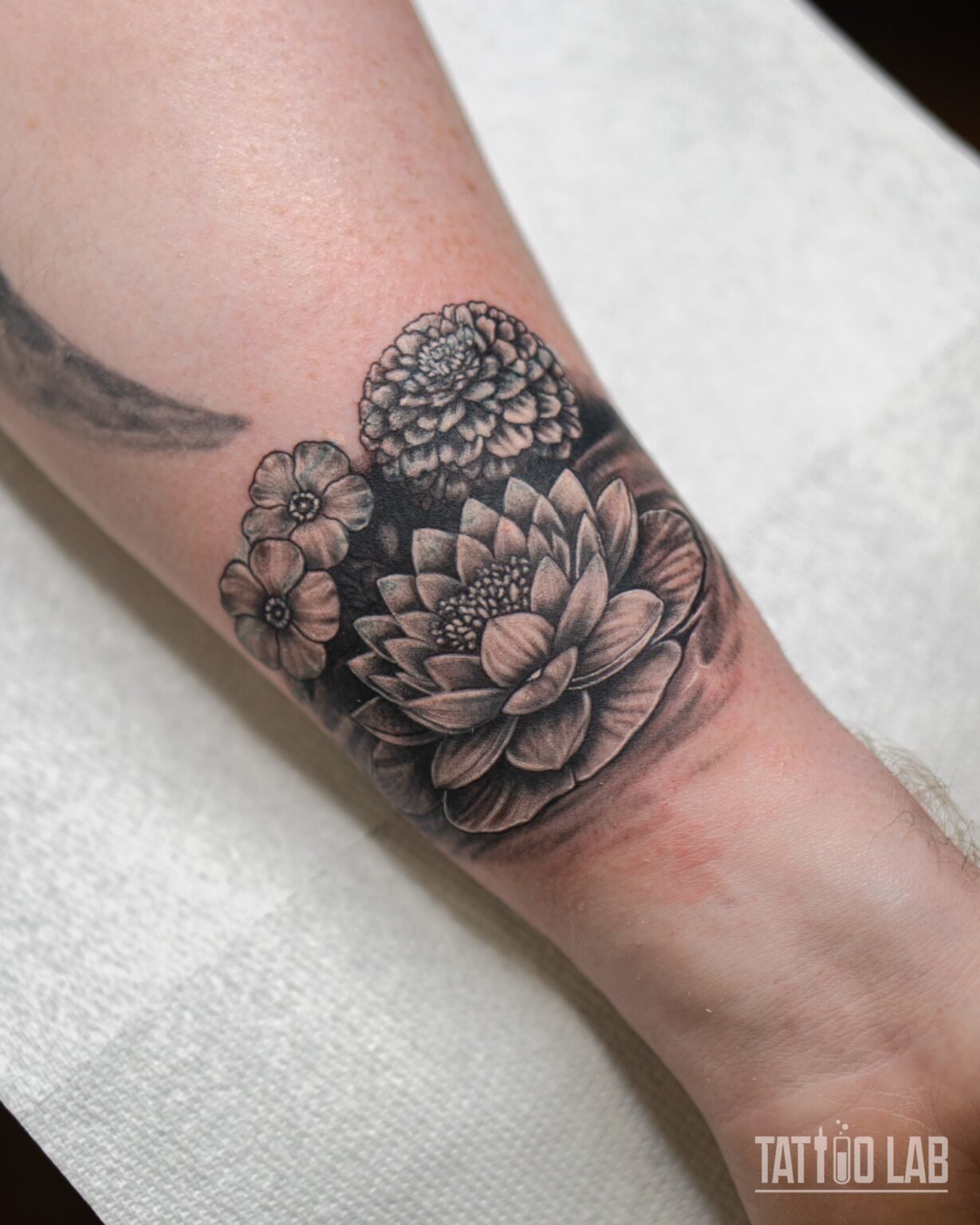 Black and grey realism cover-up tattoo of a lotus, a dahlia and small blossoms across the wrist, Tattoo Lab, Halifax