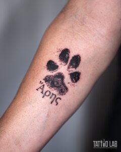 Small paw print tattoo with a stippled ink-stamp texture and short Greek lettering on the inner forearm