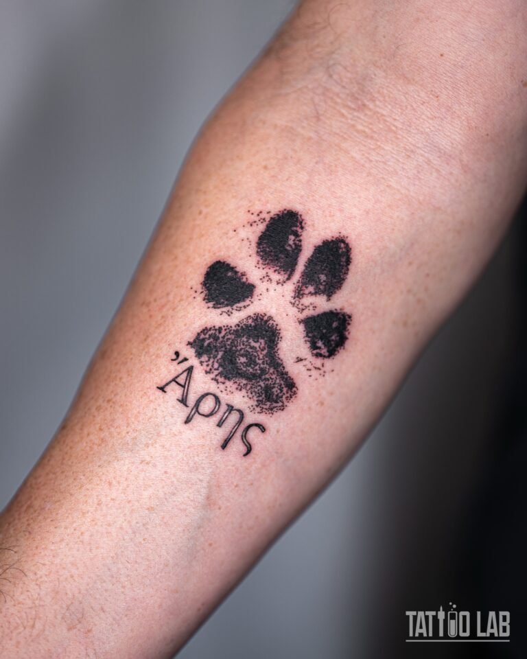 Small paw print tattoo with a stippled ink-stamp texture and short Greek lettering on the inner forearm