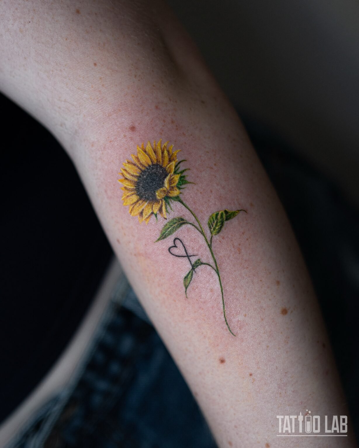 Small colour sunflower tattoo with a long green stem, two leaves and a fine line heart and infinity symbol on the inner forearm, Tattoo Lab, Halifax