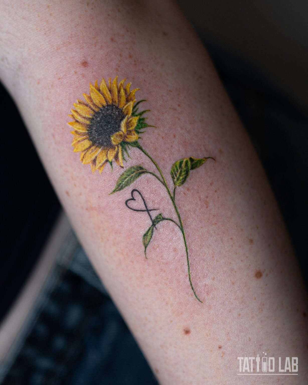 Close-up of a colour sunflower tattoo with yellow petals, a dark seed centre and a fine line heart and infinity symbol on the stem, Tattoo Lab, Halifax